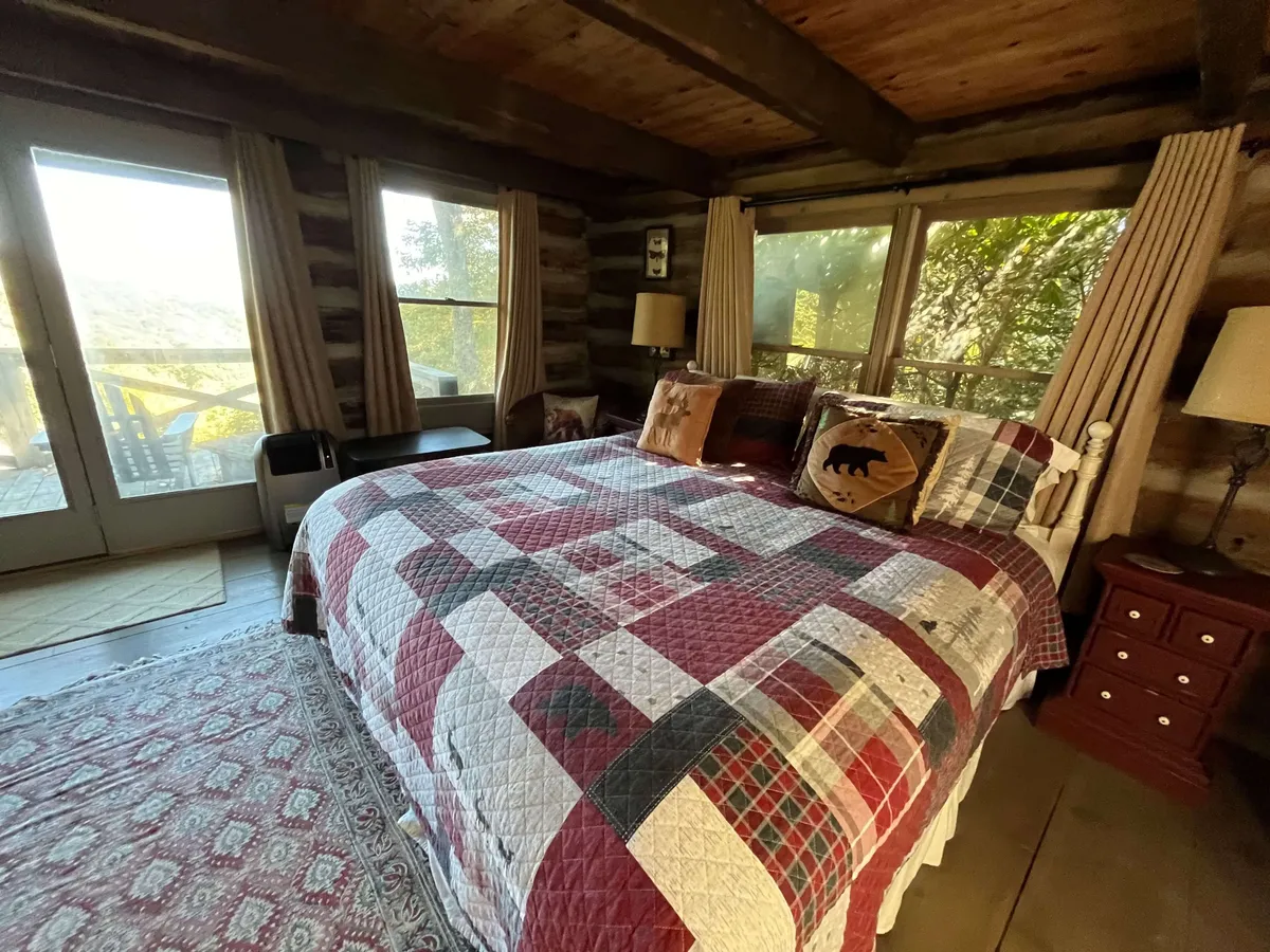 A cozy bedroom featuring rustic log walls, a plaid quilted bedspread, large windows allowing in natural light, wooden beams overhead, and decorative pillows. The windows offer tranquil views of the surrounding landscape, and the bed is invitingly made for a restful night.
