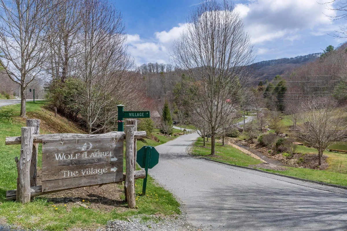 A weathered wooden sign stands alongside a quiet country roadway, framed by leafy green trees and a scenic mountain backdrop. The rustic elements suggest a longstanding presence and connection to the area’s history, inviting visitors to explore the village beyond.