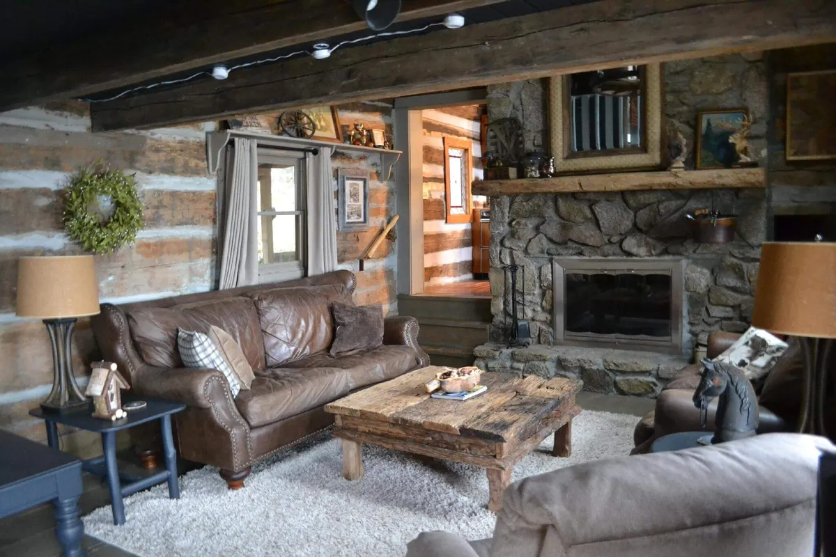 This image showcases a welcoming living room anchored by a hand-laid stone fireplace, complemented by leather sofas, a wooden coffee table, and rustic decor. Exposed wooden beams and 100+-year-old timbers are visible, infusing the area with both warmth and heritage character.
