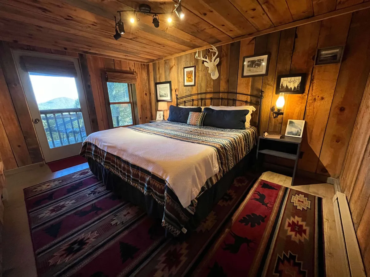 A bedroom with wooden walls, a comfortable bed topped with a warm quilt, decorative antlers above the headboard, a patterned rug, and a window revealing views to the outside. The space feels warm and deeply inviting.