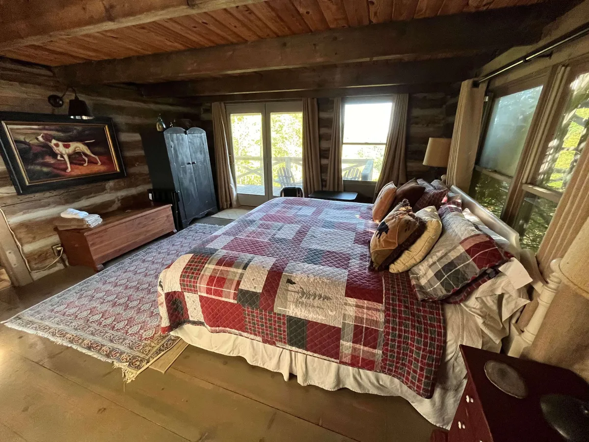 A warmly lit bedroom with a bed and quilt, large windows letting in natural light, a painting of a dog, and a wooden wardrobe. The space is designed for curling up with a book or enjoying the morning sun.