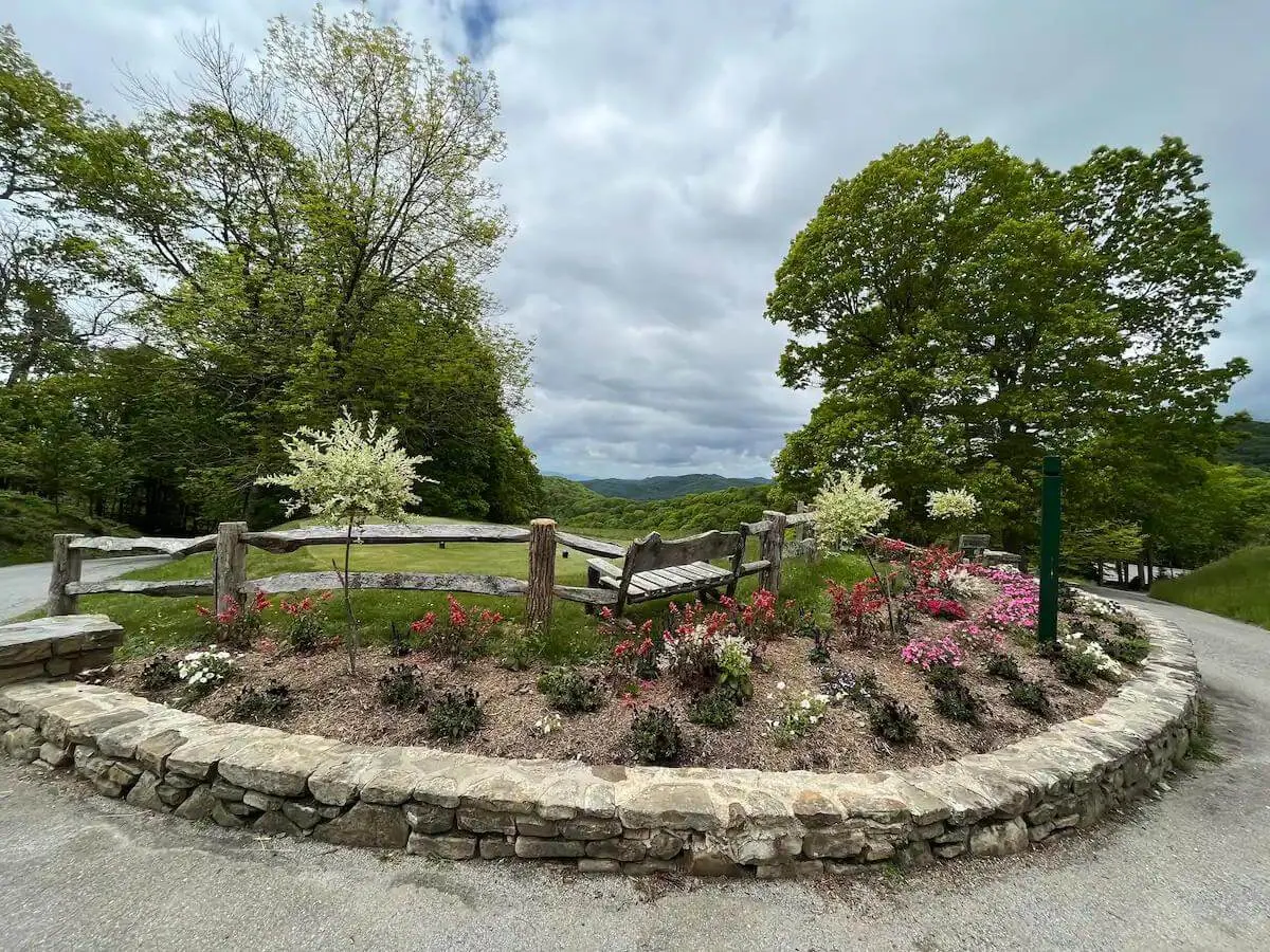 A stone flower bed and a wooden bench are nestled beneath mature trees, creating a peaceful, timeless outdoor space. The natural elements and handcrafted stonework highlight the rustic charm and enduring appeal of traditional craftsmanship.