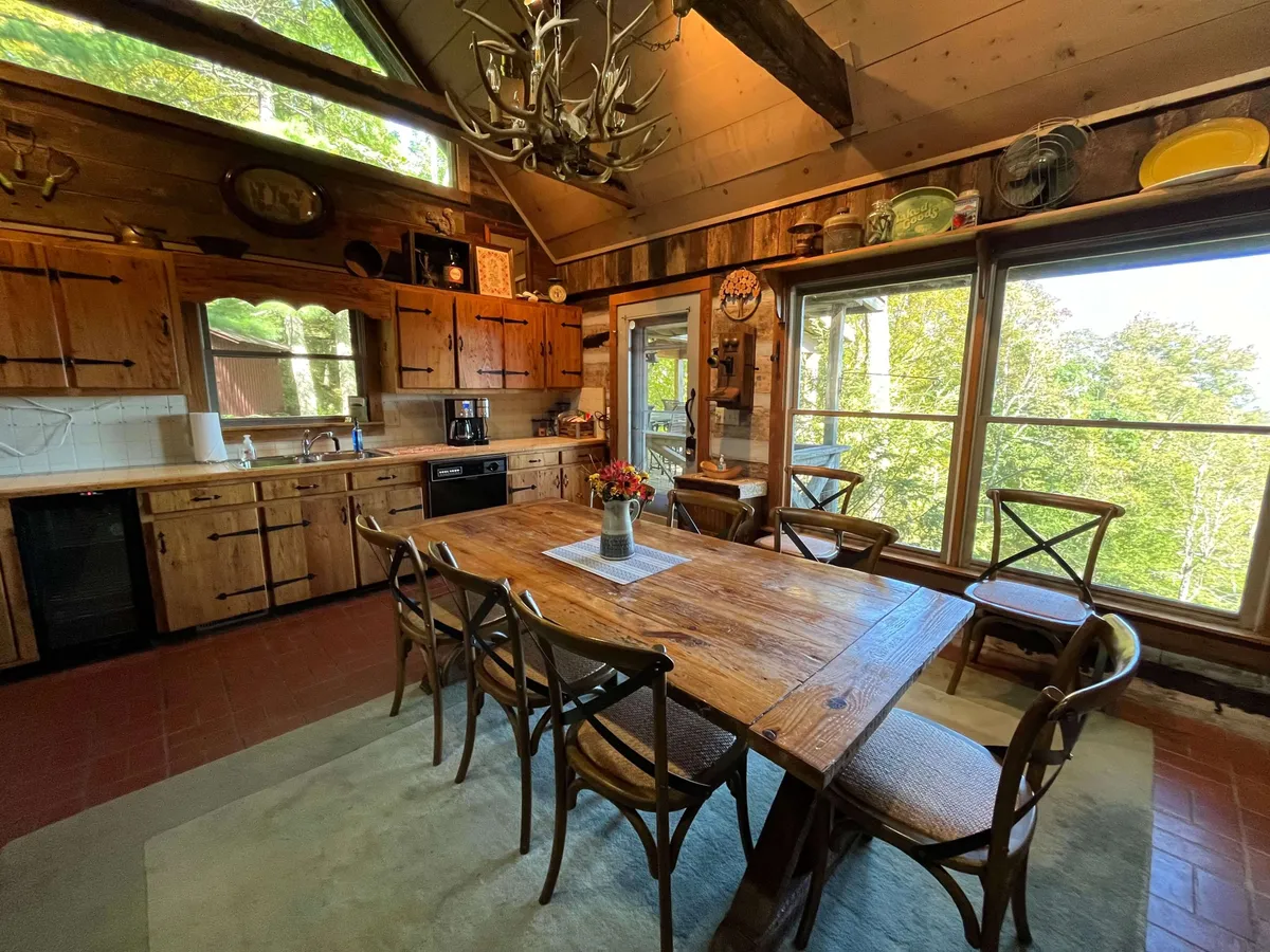 This image captures a spacious, rustic kitchen and dining area featuring a large wooden dining table, antler chandelier, and prominent kitchen cabinetry. Large windows let in natural light and offer views of the surrounding nature, while the setup invites family gatherings and social connection.